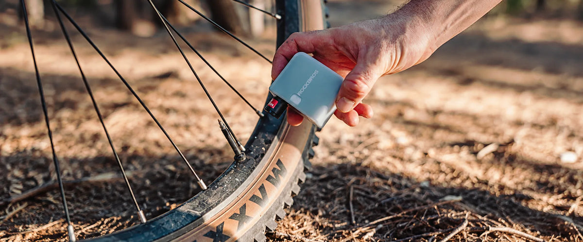 Cyclist holding a compact ROCKBROS electric bike inflator beside a mountain bike wheel on an outdoor trail